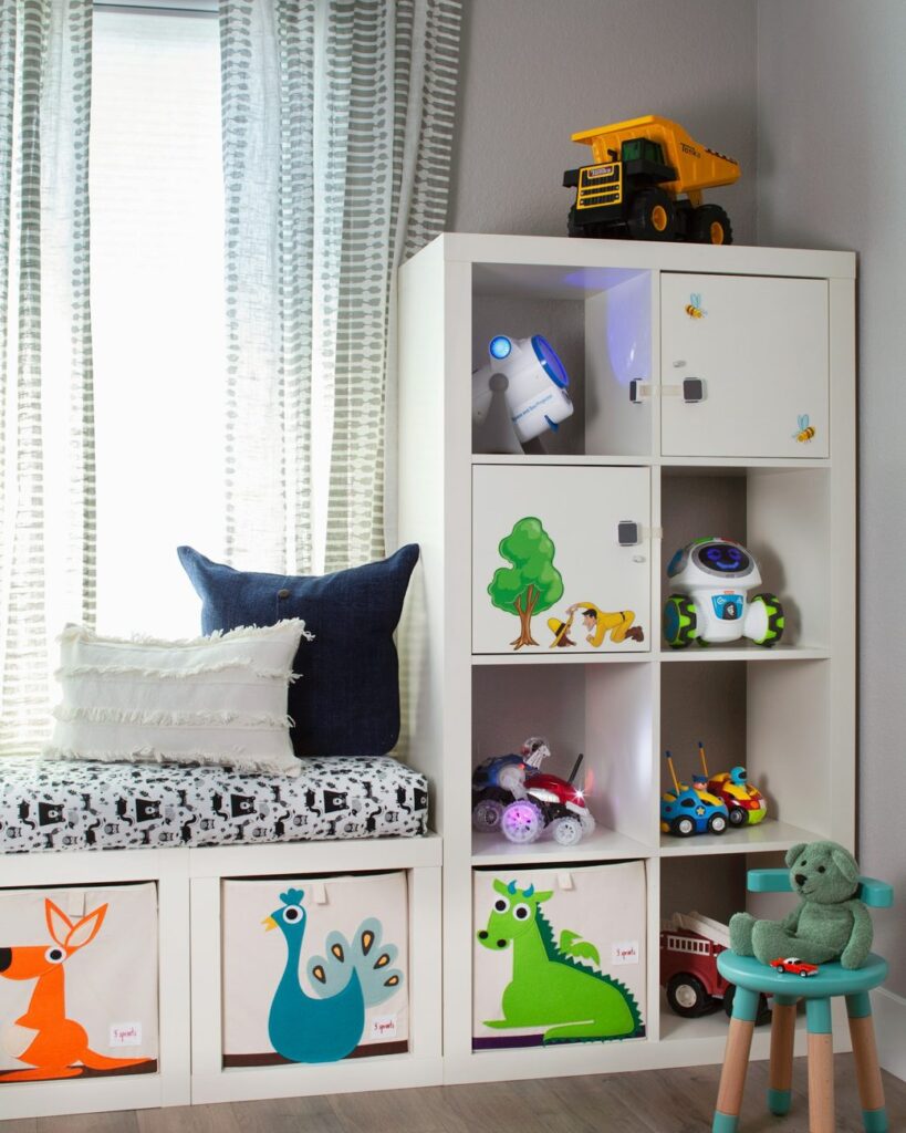 A bright playroom with a white cube shelf, toy storage boxes and a cushioned window seat